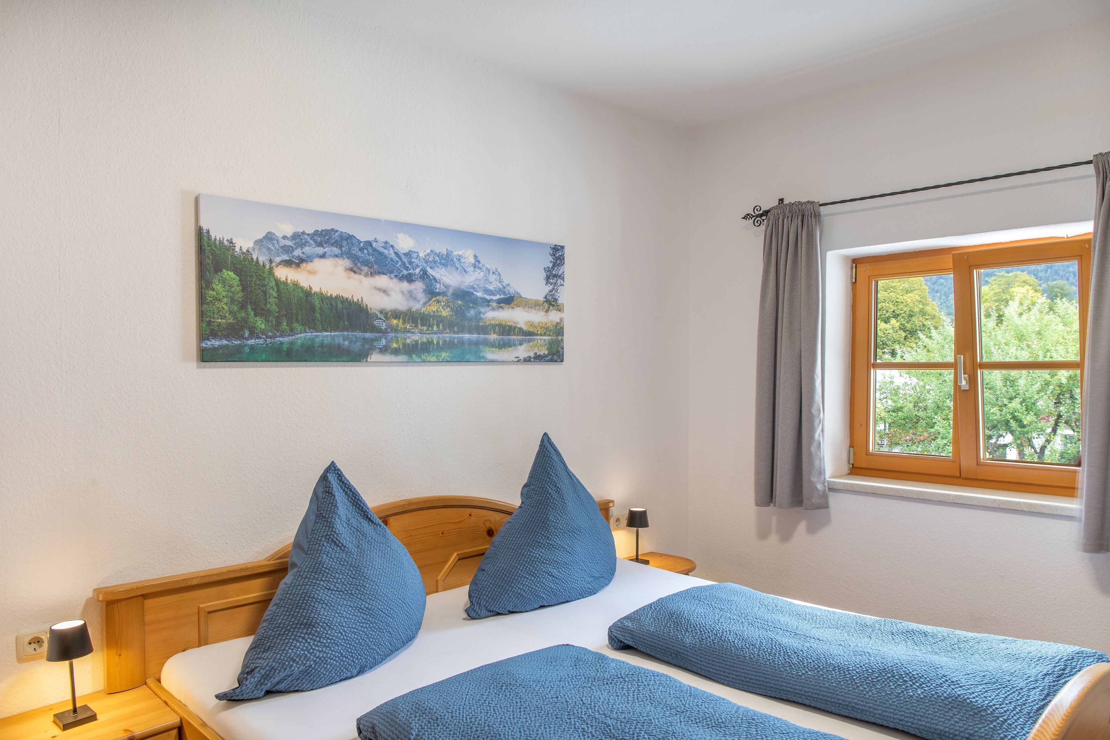 Ein Schlafzimmer mit einem Bett, Kissen, einem Nachttisch, einer Lampe, einem Fenster mit Vorhängen und einem Gemälde an der Wand.