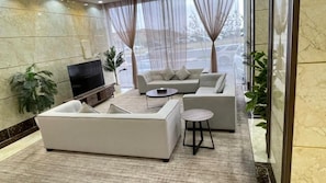 Una sala de estar moderna con un sofá modular, una mesa de centro y un televisor de pantalla plana. Hay dos plantas en macetas y un ventanal con cortinas de gasa.