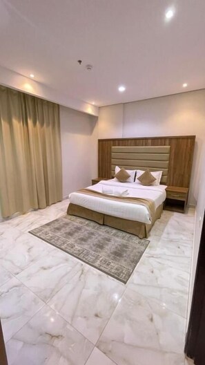 Habitación de hotel con una cama grande, cabecera de madera, piso de mármol y una cortina a la izquierda.