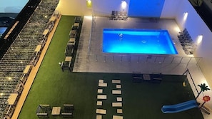Una piscina con tobogán, rodeada de zonas de descanso y un césped.