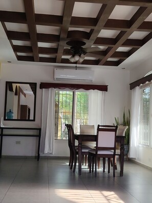 A dining area with a ceiling fan, an air conditioning unit, a dining table with chairs, and a window with curtains. - Connection, fun, relaxation—Villas Flores: a spacious home to enjoy. (TELA)