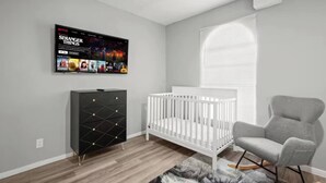 Ein moderner Babyraum mit Babybett, grauem Sessel, schwarzem Stauraum und an der Wand montiertem Fernseher, der 'Stranger Things' zeigt.
