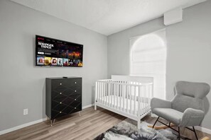 A modern baby room with a crib, a grey chair, a black storage unit, and a wall-mounted TV displaying 'Stranger Things'. - Bright Stay • DT Austin • Pecan St. Festival Ready (Austin)