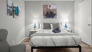 Ein modernes Schlafzimmer mit einem großen Bett, zwei Nachttischen mit Lampen, einem Sessel und Kunstwerken an den Wänden.