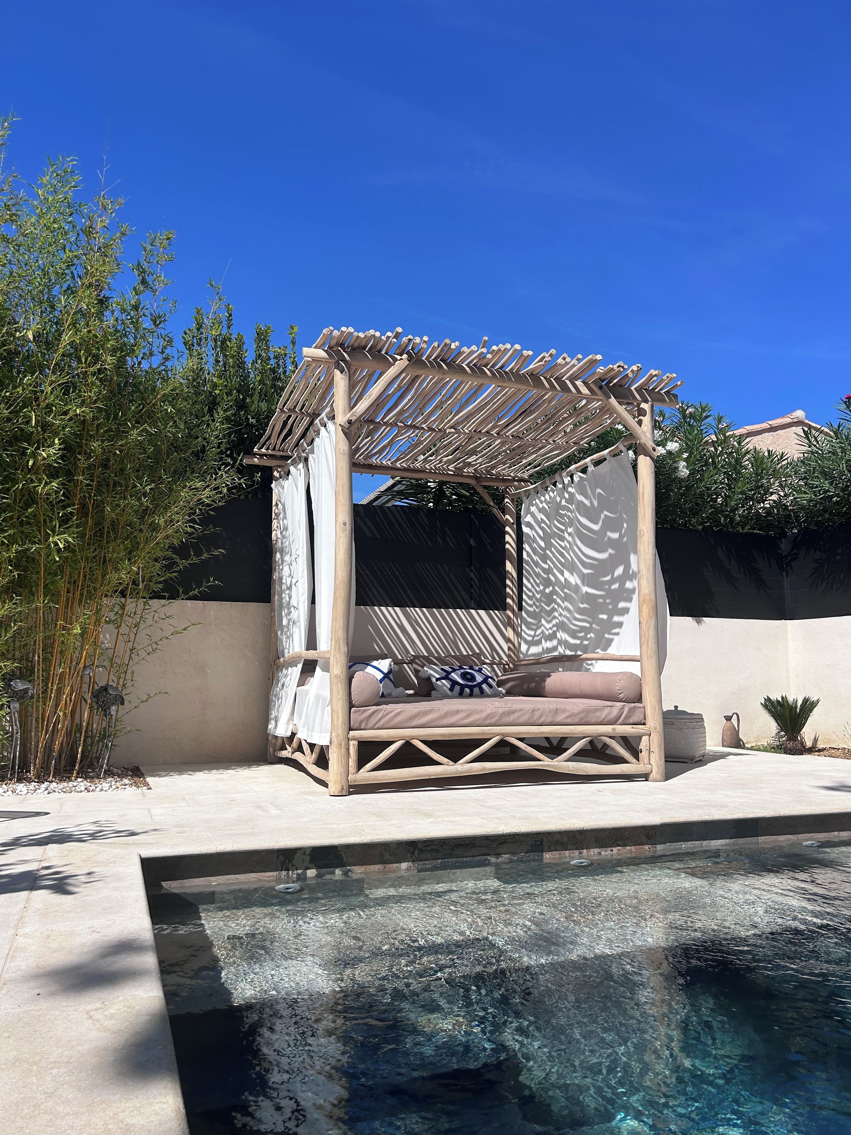 Een houten pergola met een zithoek met kussens, een zwembad en omliggende beplanting.