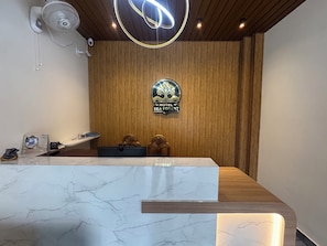 A hotel reception desk with a wooden panel wall, a logo, and a ceiling light fixture.