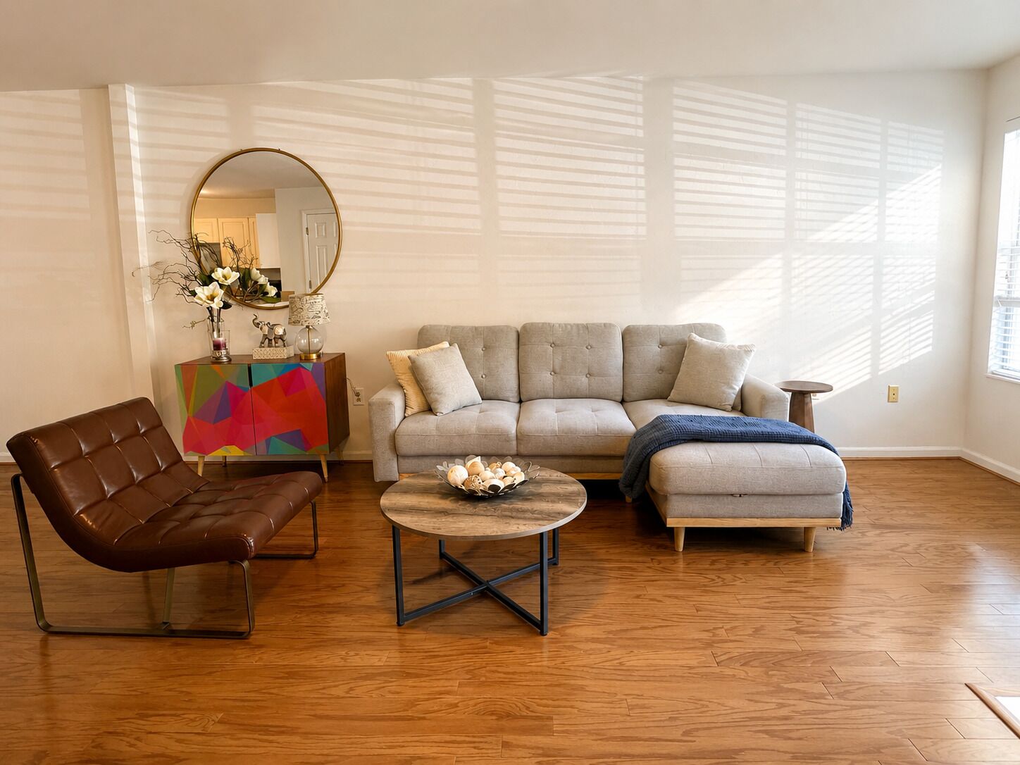 A living room with a sofa, a coffee table, and a leather chair. There is a colorful cabinet and a mirror on the wall.