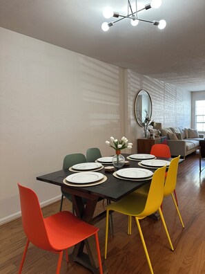 Un coin repas avec une table noire dressée pour six personnes, entourée de chaises colorées, un miroir au mur et un canapé à l’arrière-plan.