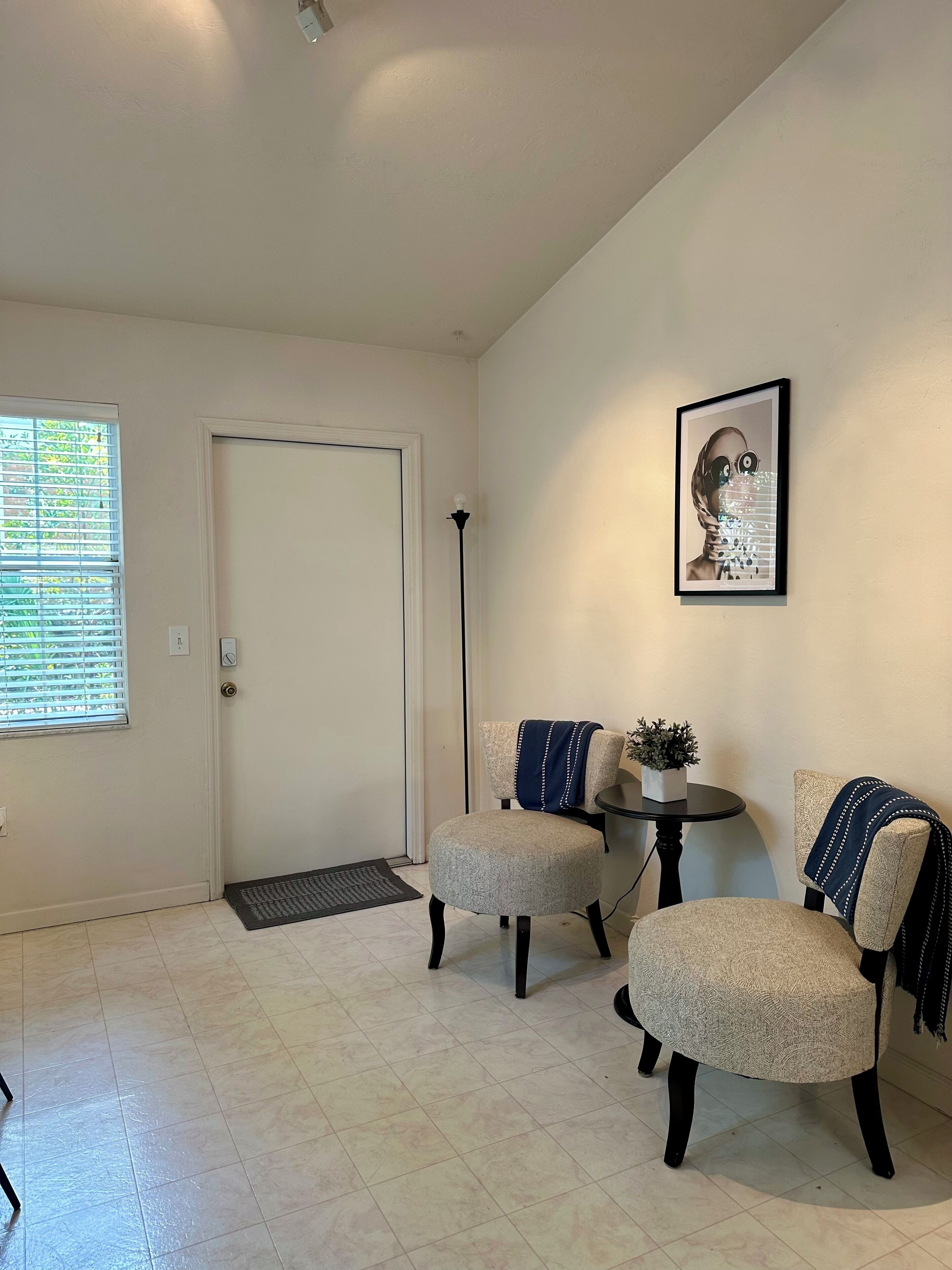 Ein Zimmer mit zwei Polstersesseln, einem kleinen runden Tisch, einem gerahmten Bild an der Wand und einer Tür mit Fenster und Jalousien.