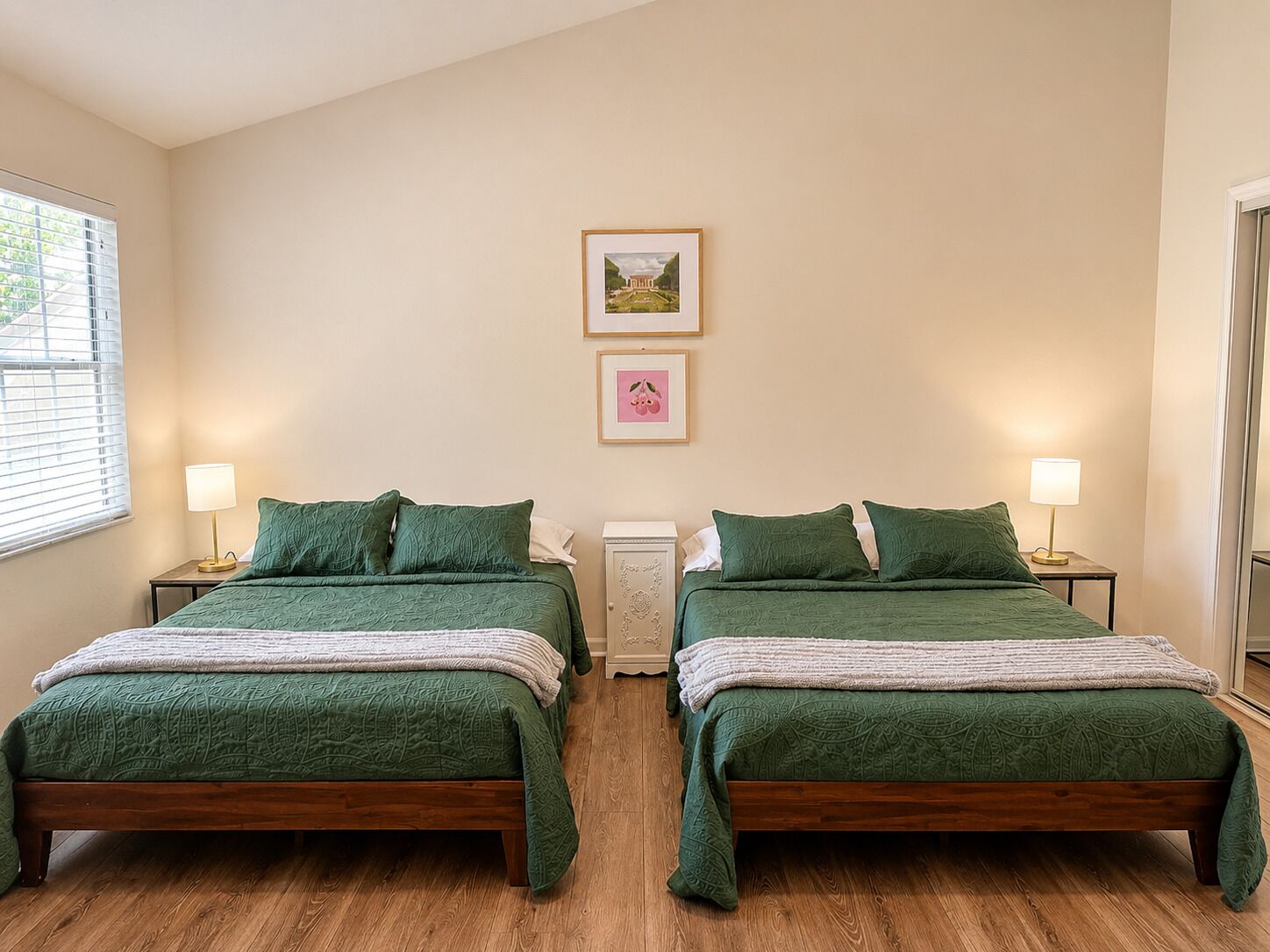 Una habitació amb dos llits, cadascun amb un estovat i coixins verds, flanquejats per taules de nit amb làmpades. Al mur, sobre els llits, hi ha un quadre amb marc.