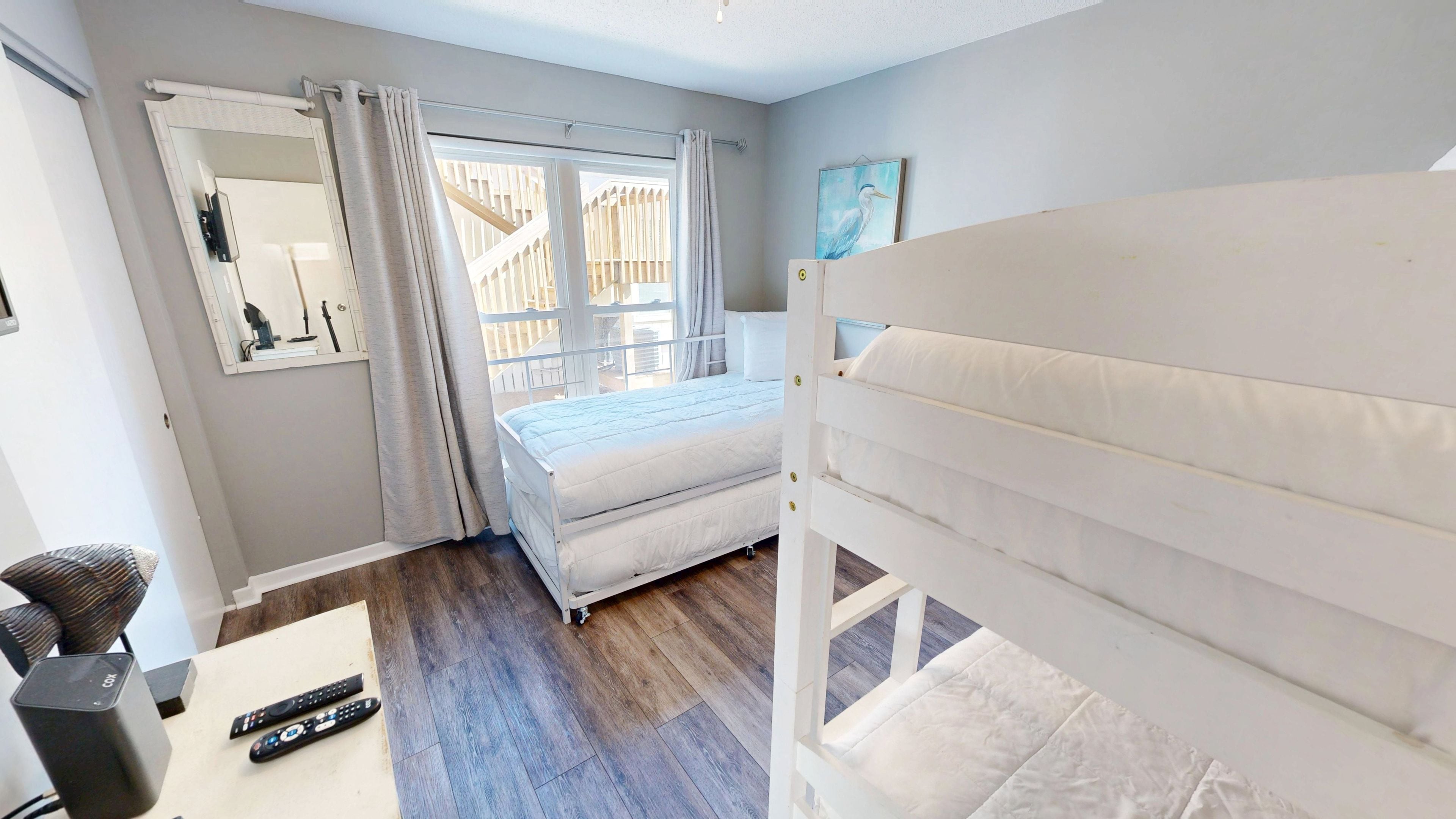 A bedroom with a bed, a chair, a table with a speaker and remote controls, a window with curtains, and a painting on the wall.