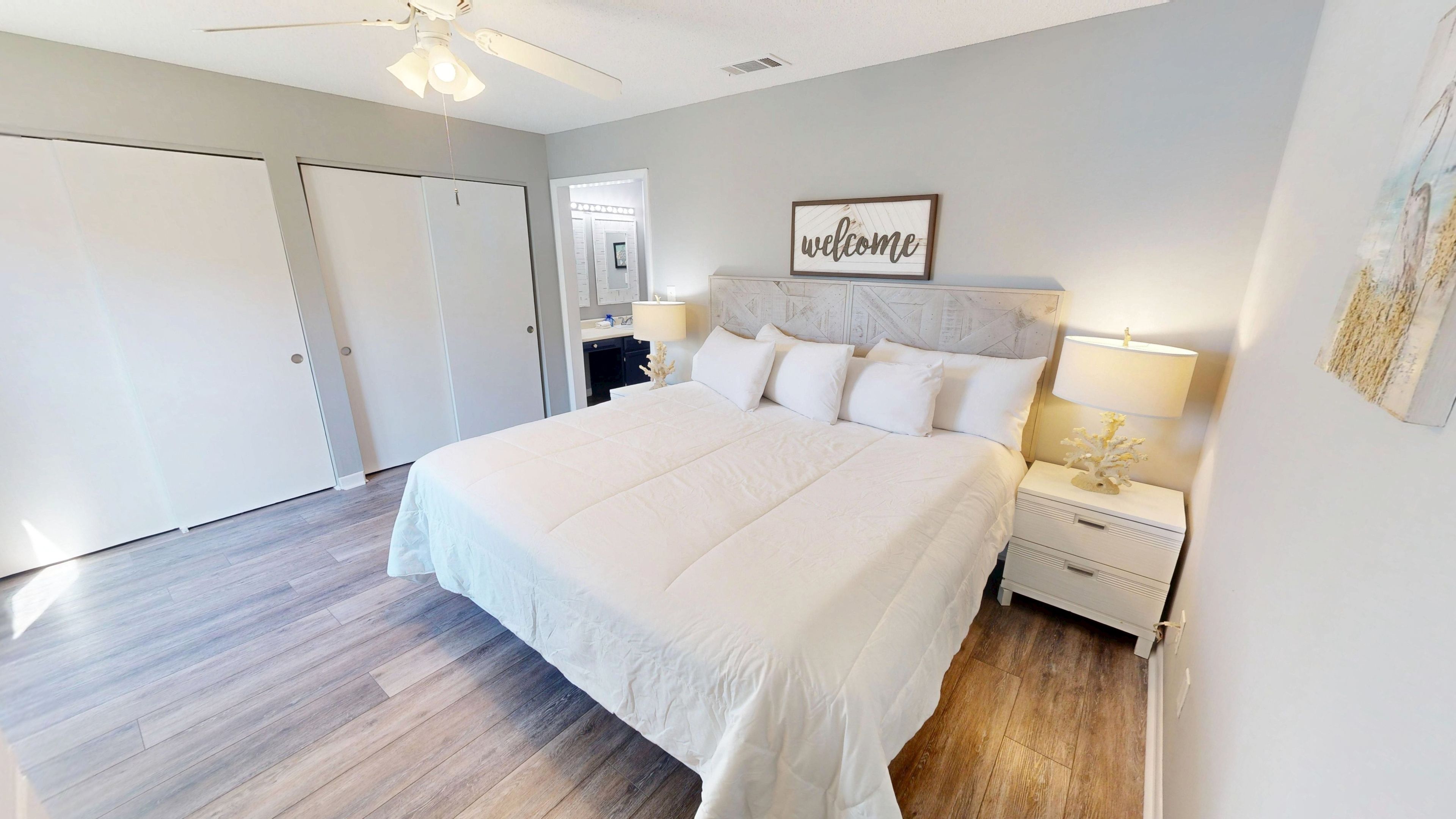 A bedroom with a large bed, a nightstand, a wall-mounted lamp, a ceiling fan, and a door.