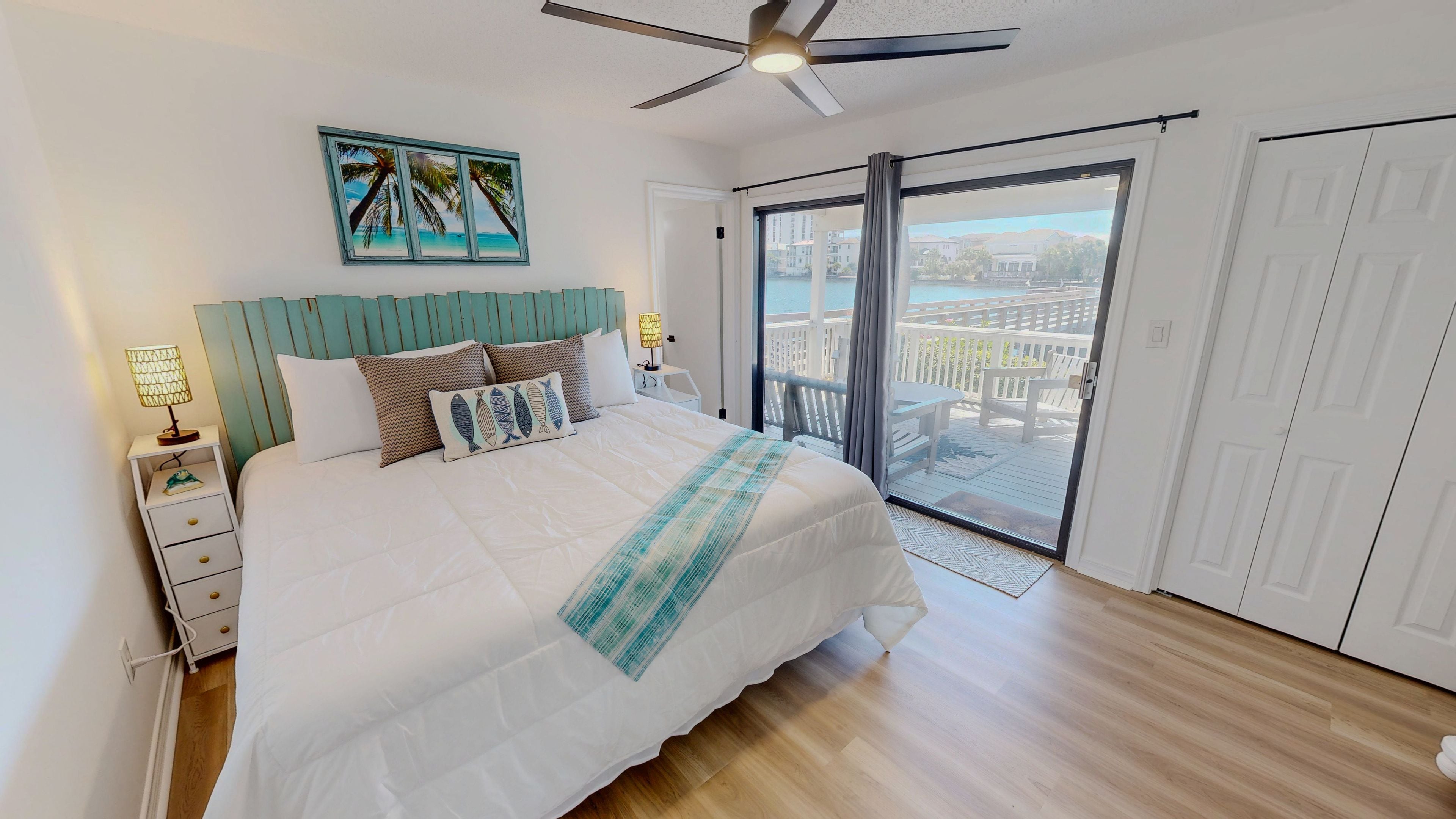 A bedroom with a bed, a nightstand, a ceiling fan, a window with a view of a balcony, and a door.