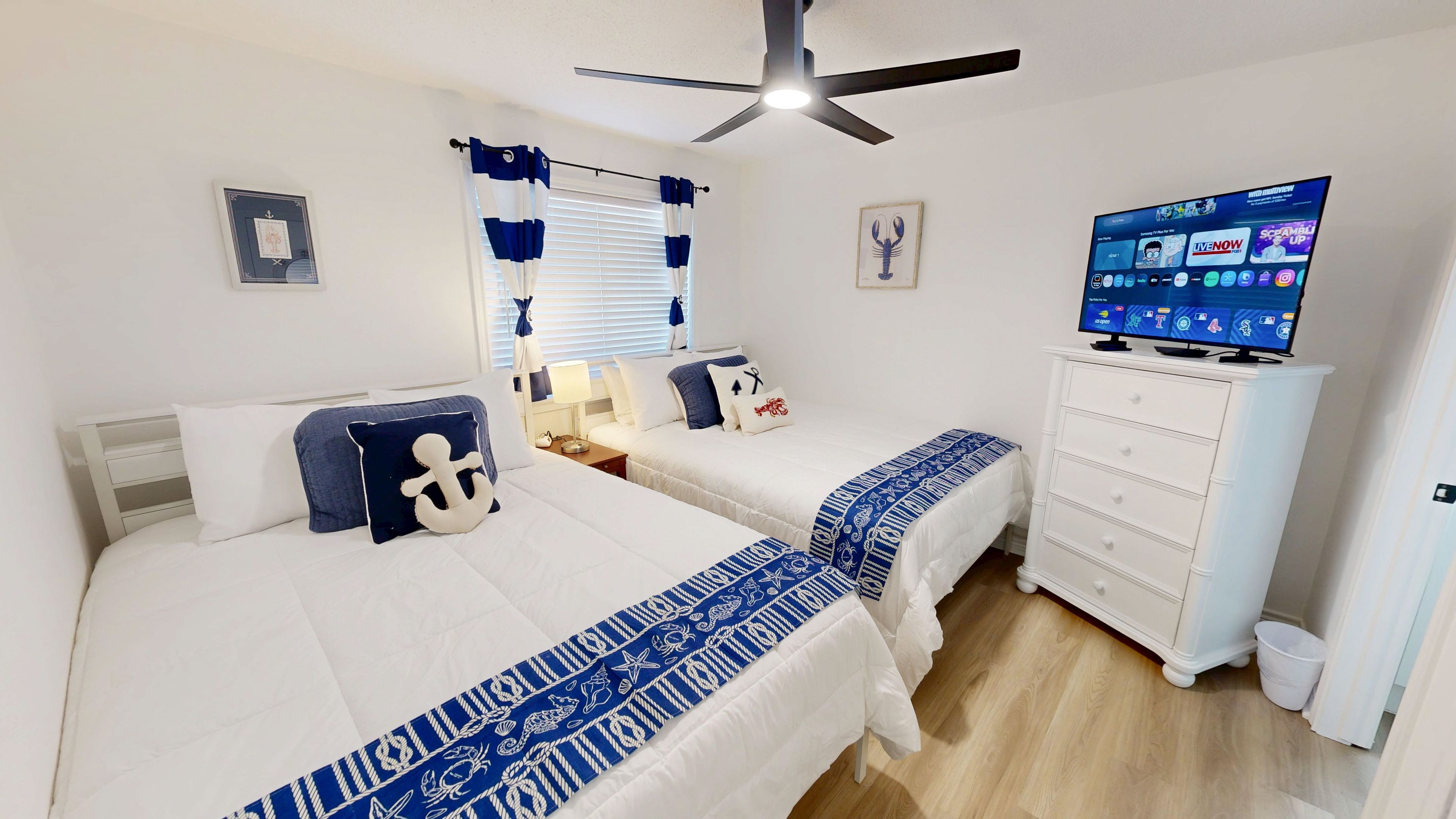Une chambre avec deux lits, un ventilateur de plafond, une télévision et une commode.
