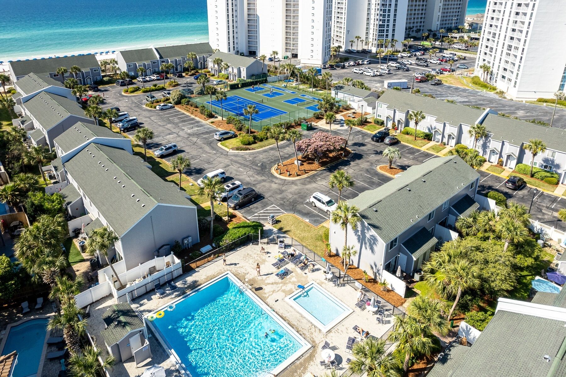 Vista aérea de un hotel con varias construcciones, estacionamiento, cancha de tenis, piscina y vistas a la playa.