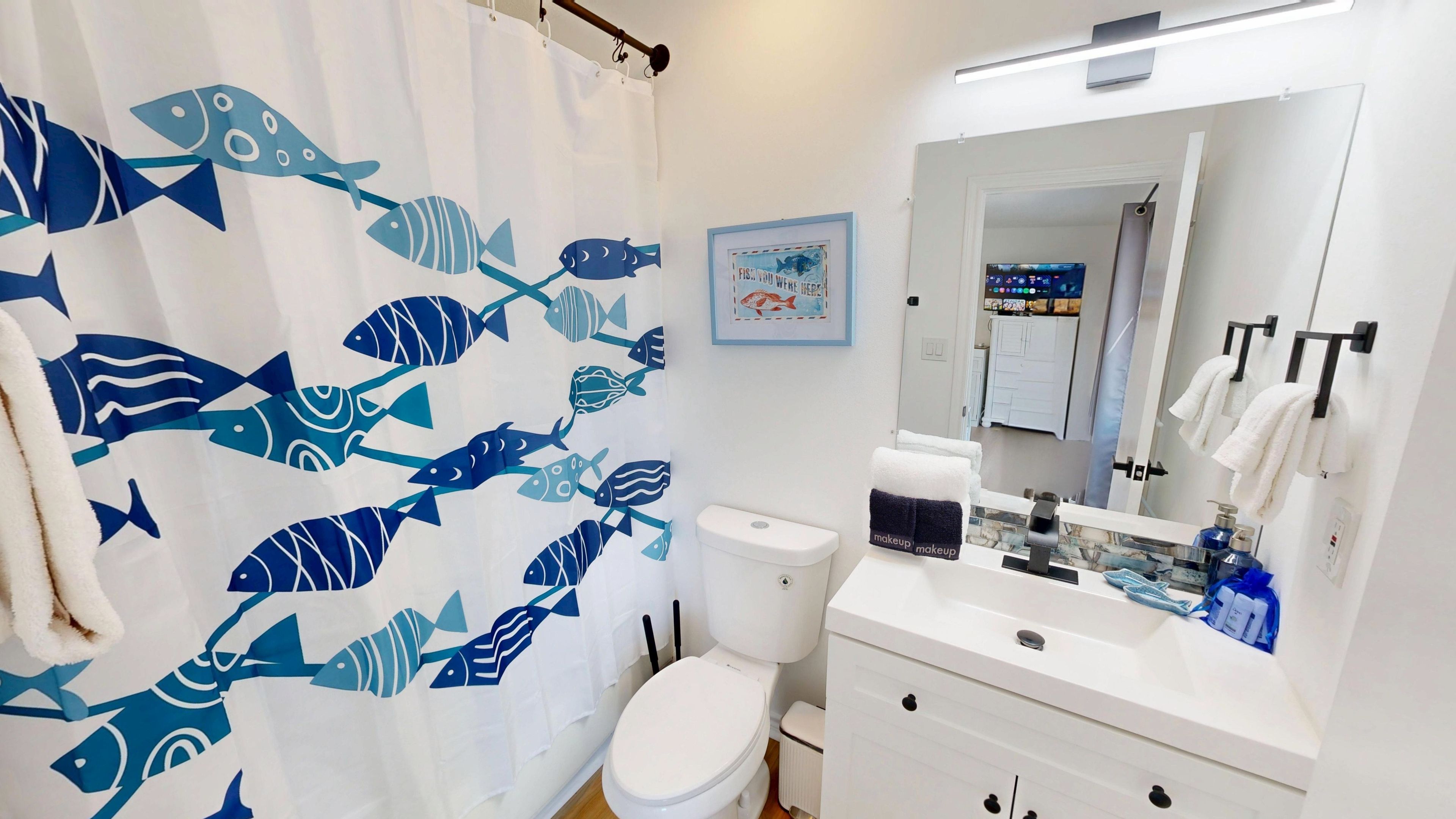Un bagno con tenda da doccia a motivo di pesce, un water, un lavandino con specchio e un quadro appeso al muro.