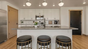 Uma cozinha moderna com armários brancos, uma geladeira de aço inoxidável, uma ilha central com banquetas e iluminação pendente.