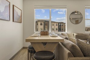 A modern living room with a dining area, a large window with a view of buildings, and a round mirror on the wall. - Adventure Loft 3 Bedroom Condo (Hurricane)
