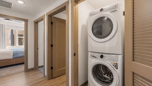 Uma lavanderia com máquina de lavar e secadora empilhadas, piso de madeira e uma cama no quarto adjacente.