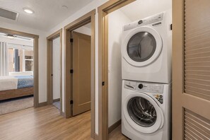 Ein Waschraum mit einer gestapelten Waschmaschine und einem Trockner, Holzboden und einem Bett im angrenzenden Zimmer.