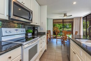 A modern kitchen with a built-in oven, microwave, and coffee maker. There is a dining area with a table and chairs, and a view of greenery outside. - Wailea Ekahi 3a 2 Bedroom Condo (Kihei)