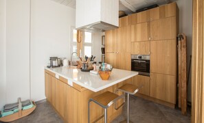 Eine moderne Küche mit Holzschränken, einer zentralen Kochinsel mit Barhocker, einem eingebauten Backofen und verschiedenen Küchenutensilien.