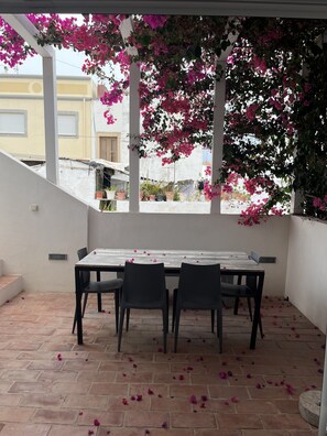 A covered patio with a table and chairs, surrounded by potted plants and pink flowers. - Holiday Home 'Casa Opala Townhouse with Pool' , Wi-Fi and Air Conditioning (Olhão)