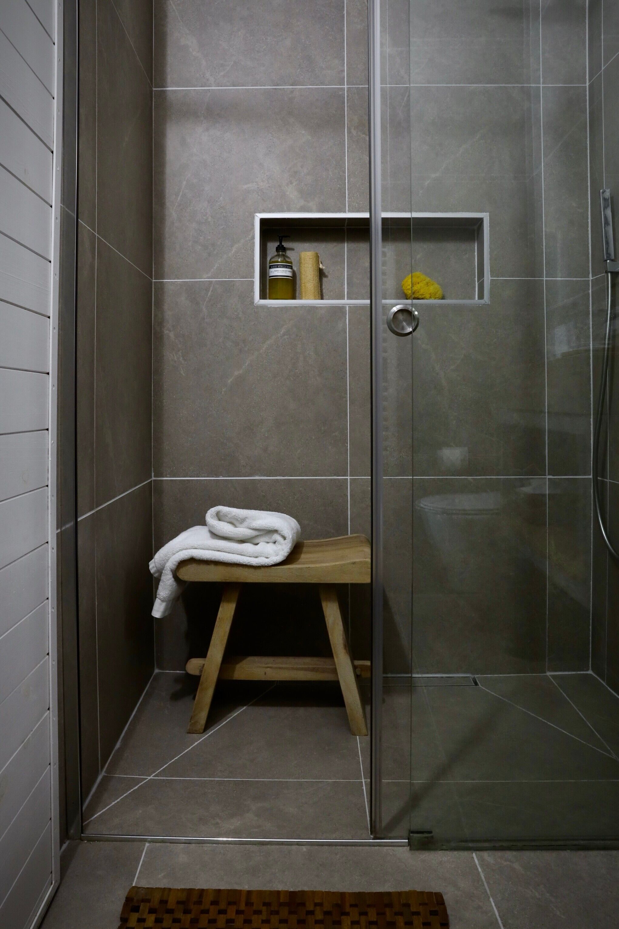 A shower area with a glass door, a wooden stool, a white towel, and a shelf with toiletries.