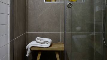 A shower area with a glass door, a wooden stool, a white towel, and a shelf with toiletries.