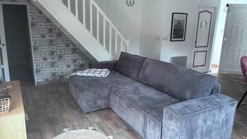 Een moderne woonkamer met een grijze hoekbank, een houten salontafel, een tapijt met een patroon, een trap met een witte balustrade en een deur met een kunstwerk erin.