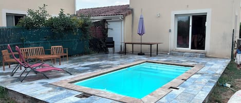 A swimming pool with a tiled deck, a table, chairs, and a covered umbrella.