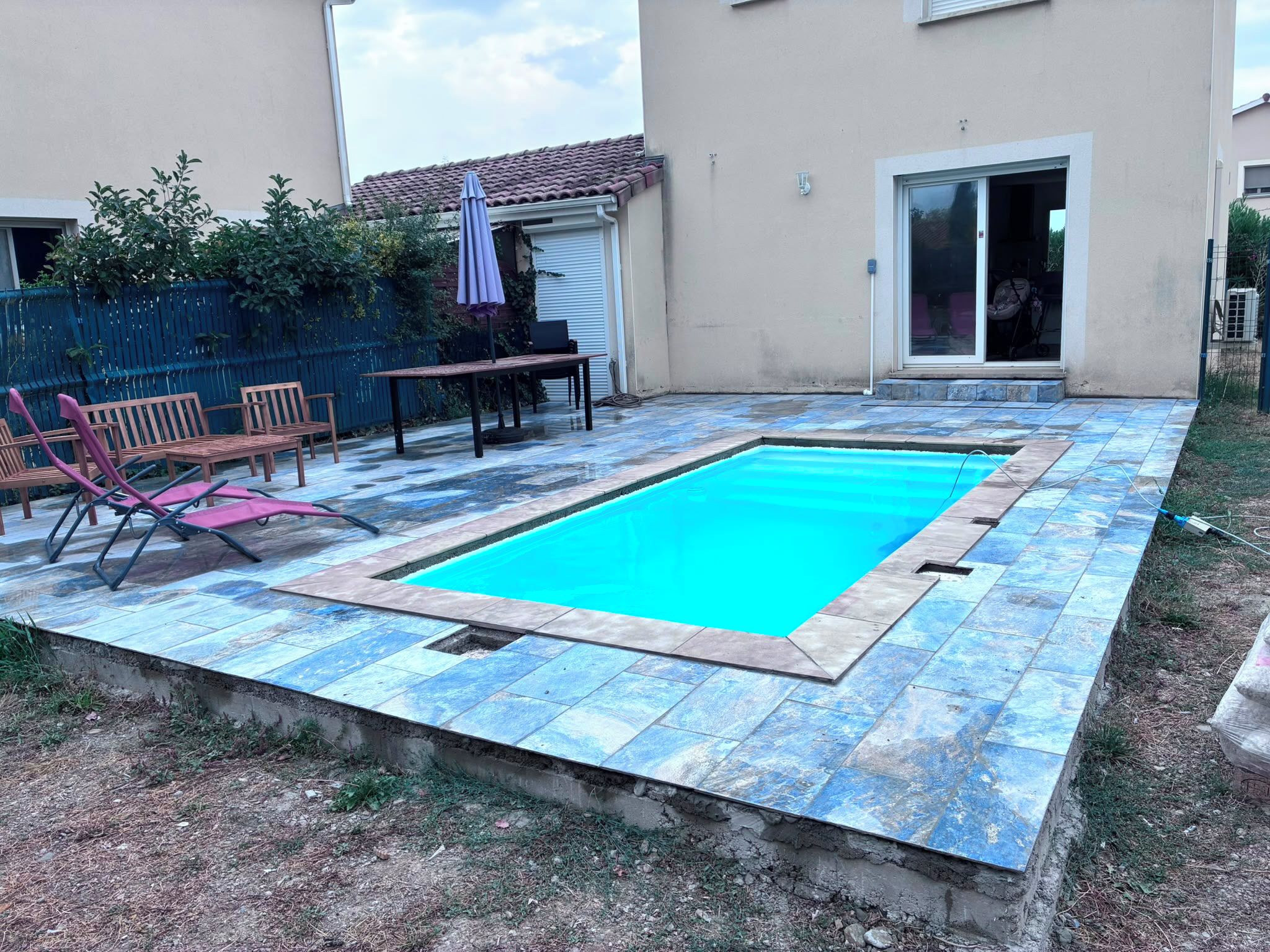 A swimming pool with a tiled deck, a table, chairs, and a covered patio area.