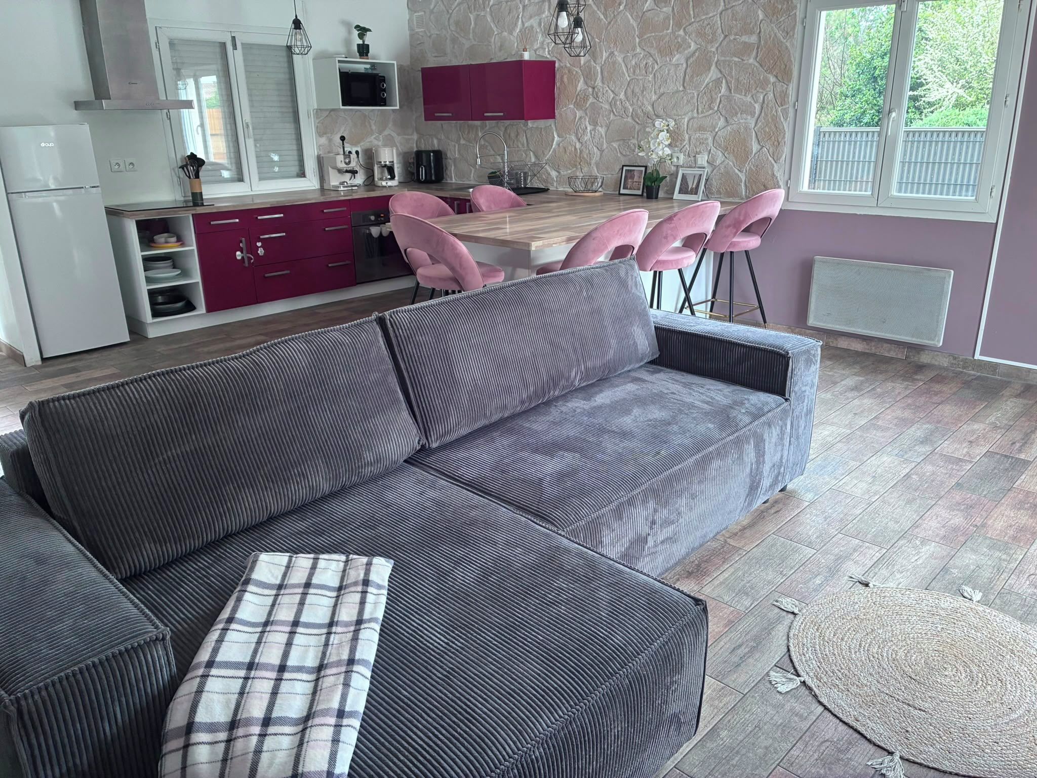 Eine moderne Küche mit einer Steinwand, einem Essbereich mit rosa Stühlen und einem grauen Sofa mit einem Karokissen.
