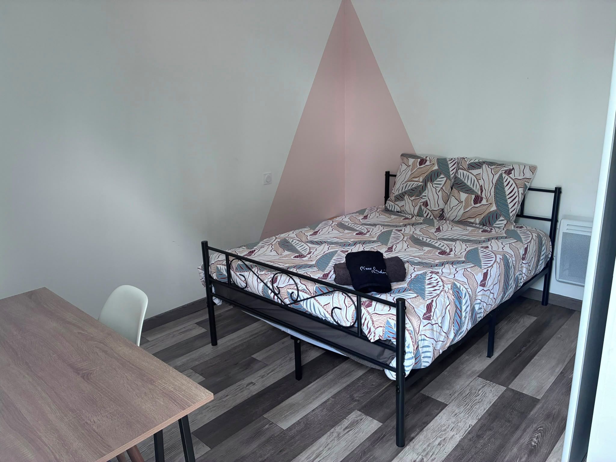 A single bed with a patterned bedspread, a small table, and a chair in a room with a sloped ceiling and a radiator.