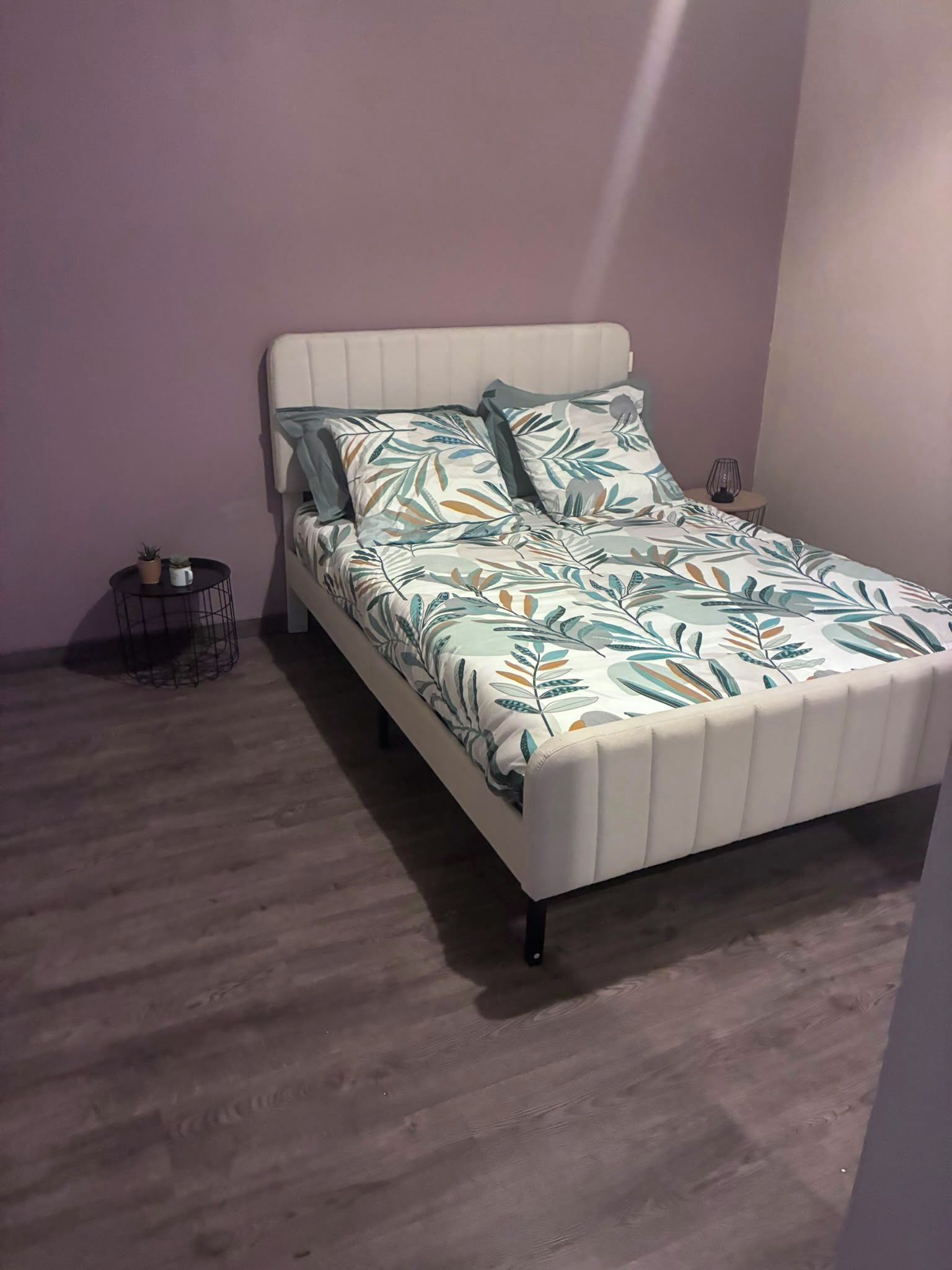 Un lit simple avec une couvre-lit à motifs de feuilles et des oreillers, une petite table d’appoint ronde avec une plante, et un petit panier en fil de fer.