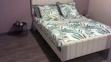 Un lit simple avec une couvre-lit à motifs de feuilles et des oreillers, une petite table d’appoint ronde avec une plante, et un petit panier en fil de fer.