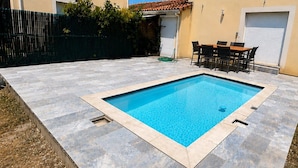 A rectangular swimming pool with a tiled deck, surrounded by a fenced area with a small patio and a table with chairs. - The Sunny House (Bérat)