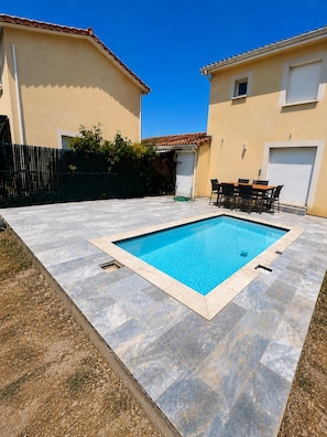Et rektangulært pool med flisebelagt solterrasse, omgivet af et hegn med en lille patio og et bord med stole.
