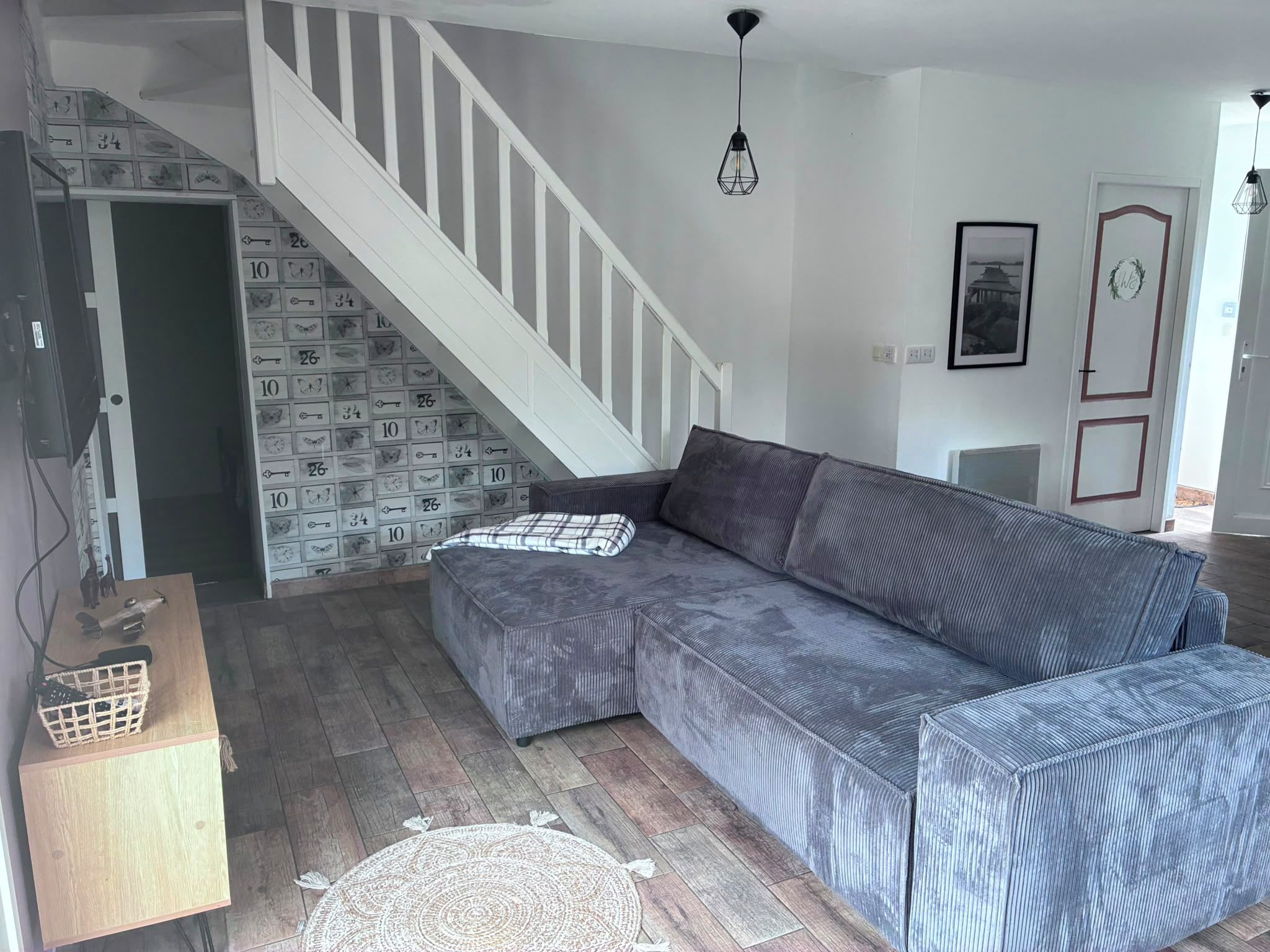 Ein Wohnzimmer mit grauem Sofa, einem Holztisch, einer Treppe mit weißer Geländer und einer Wand mit Muster.