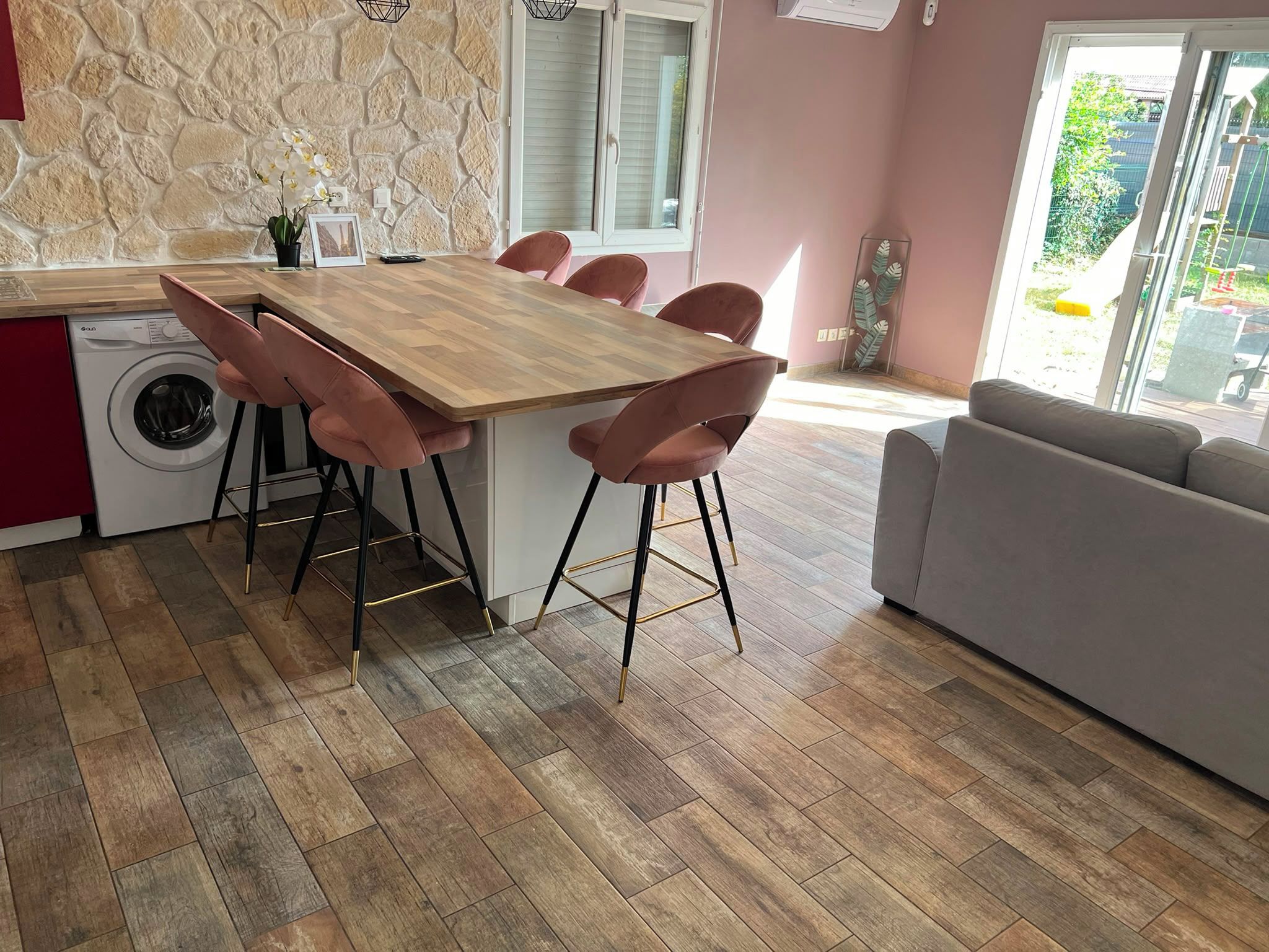Eine moderne Küche mit Holztisch und rosa Stühlen, einer Waschmaschine und einem grauen Sofa in einem hellen Raum mit Steinwand.