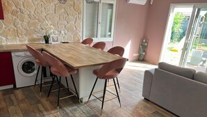 A modern kitchen with a wooden table and pink chairs, a washing machine, and a grey sofa in a bright room with a stone wall and a large window. - The Sunny House (Bérat)
