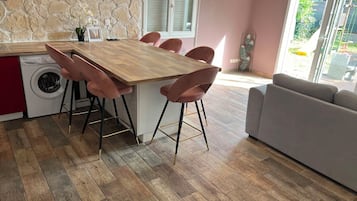 Eine moderne Küche mit Holztisch und rosa Stühlen, einer Waschmaschine und einem grauen Sofa in einem hellen Raum mit Steinwand.
