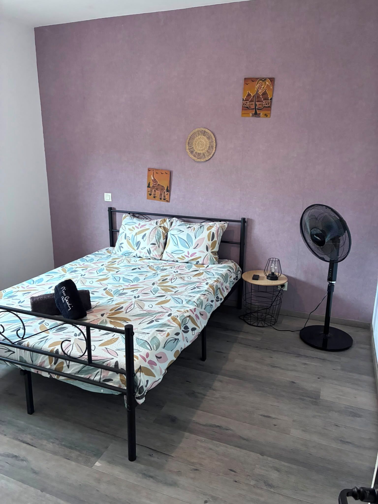 Een slaapkamer met een bed, een ventilator, een tafeltje met een lamp en twee decoraties aan de muur.