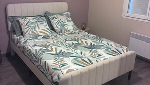 A single bed with a leaf-patterned bedspread and matching pillows, a small bedside table with a lamp, and a window with blinds. - The Sunny House (Bérat)