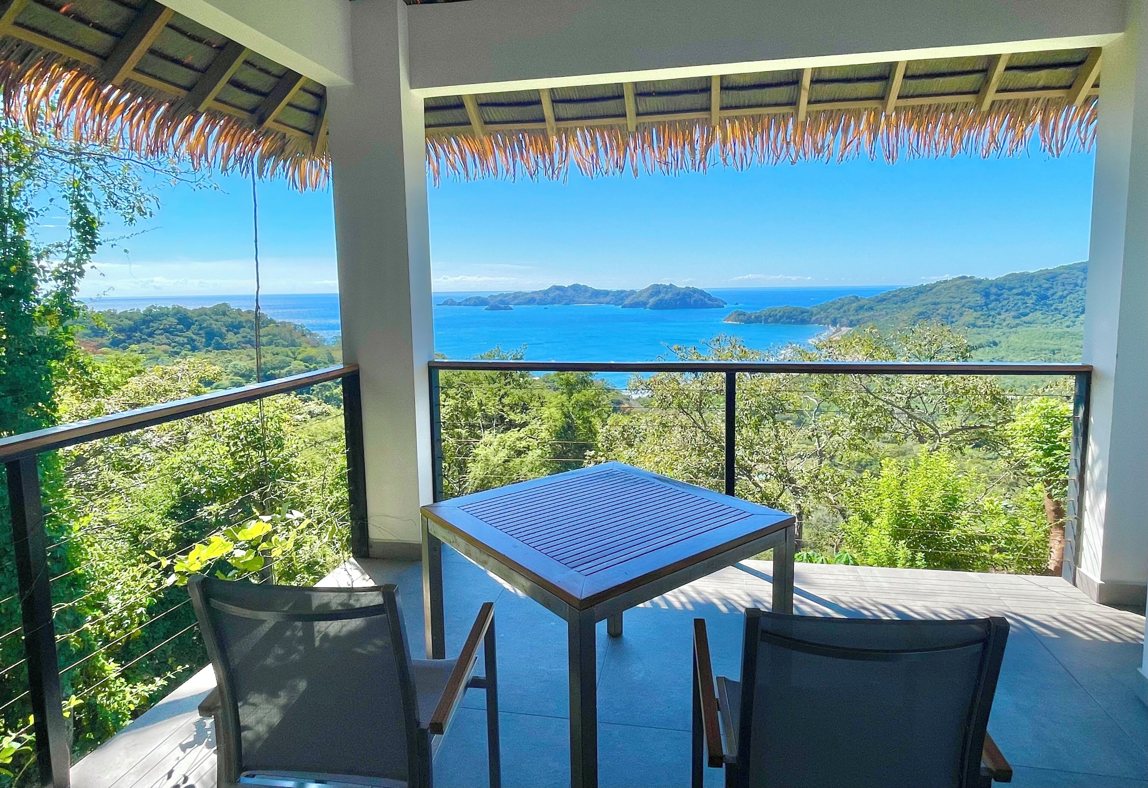 A balcony with a table and chairs overlooking a scenic view of the sea and hills.