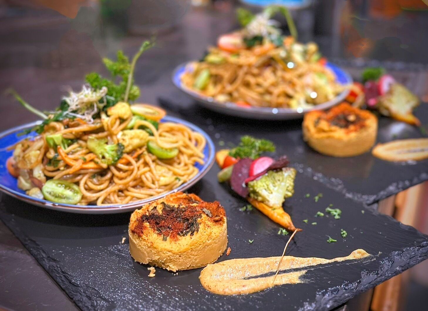 Un tavolo da pranzo con piatti di noodles e verdure, una fetta di pane e una piccola ciotola di salsa.