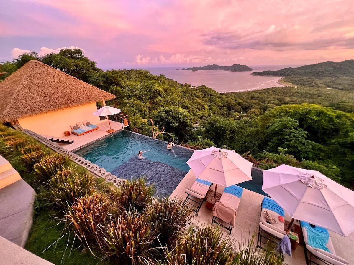 A pool area with lounge chairs and umbrellas, surrounded by lush greenery and overlooking a body of water.
