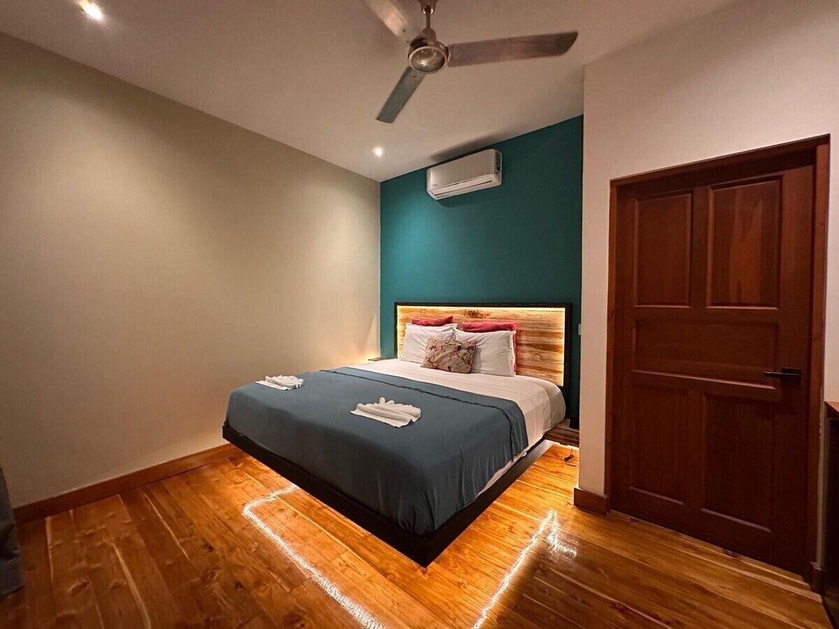 A bedroom with a bed, a ceiling fan, an air conditioning unit, and a wooden door.