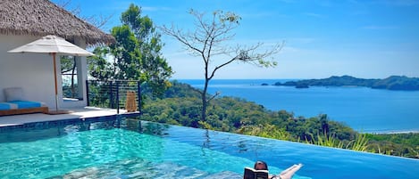Una piscina infinita con agua azul cristalina, un área de descanso con techo de paja y vistas al océano y las colinas.