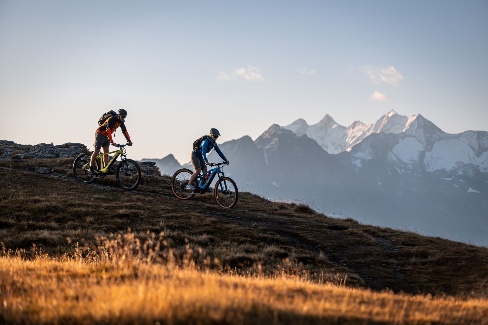 Two cyclists on a mountain trail with a view of snow-capped mountains in the background.